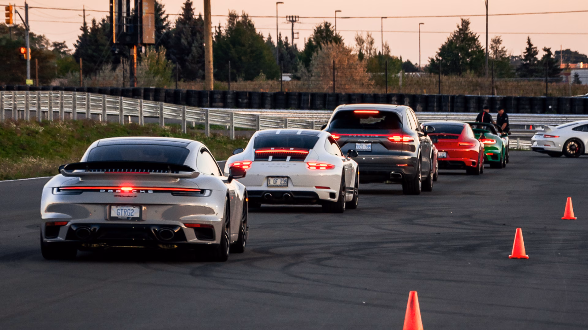 A line of Porsches driving out in convoy at dusk, marker cones along the edge of the road